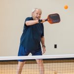 A man in a navy shirt laughs while swinging his paddle at an orange pickleball near the net.
