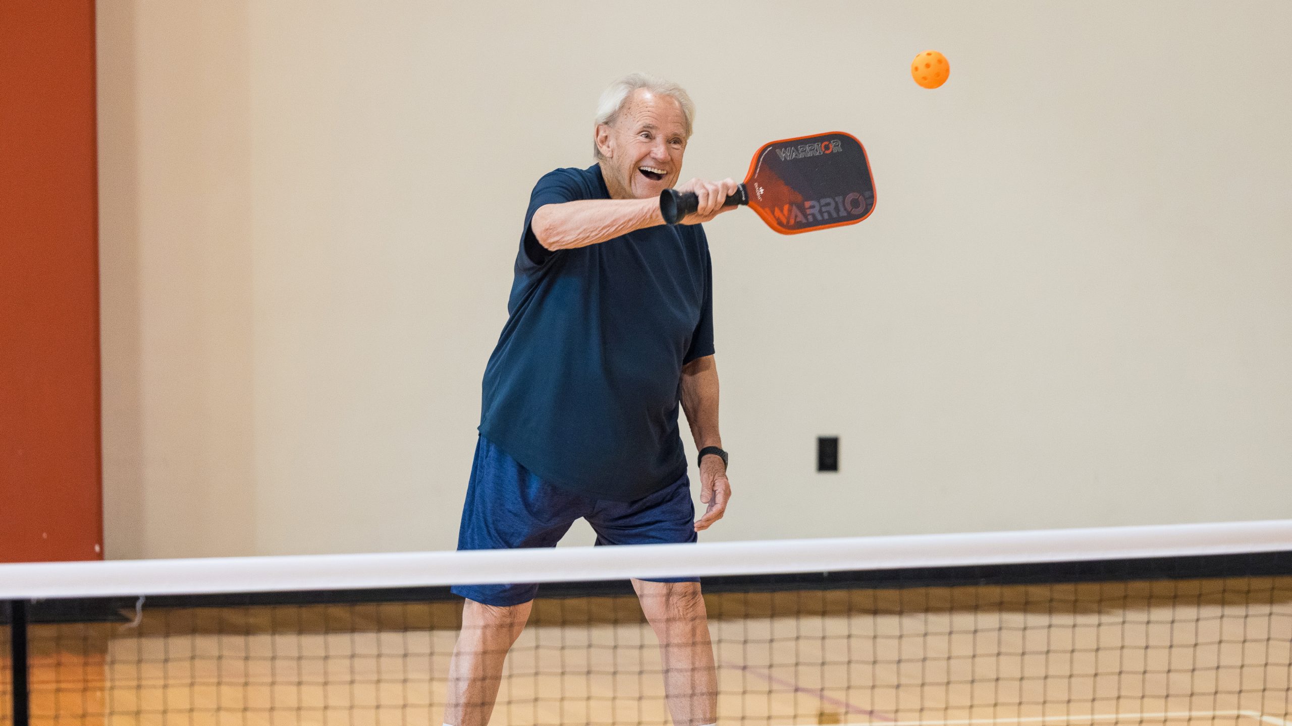 A man in a navy shirt laughs while swinging his paddle at an orange pickleball near the net.