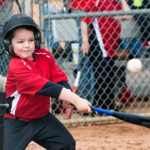 A young child wearing a helmet and red baseball uniform swings a bat at a baseball during a tee-ball game, with spectators and a chain-link fence in the background.