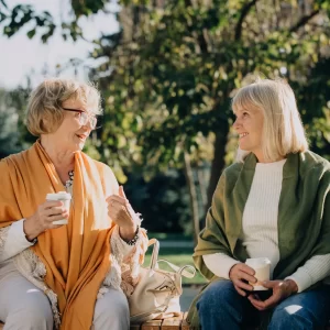 Two senior women sharing a moment of friendship, talking and drinking coffee on a park bench during a sunny autumn day