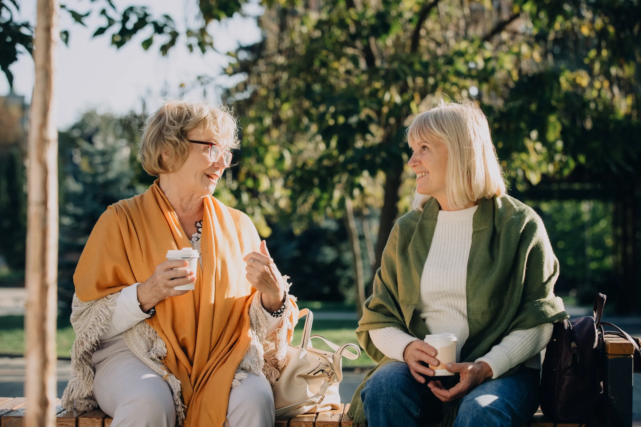 Two senior women sharing a moment of friendship, talking and drinking coffee on a park bench during a sunny autumn day